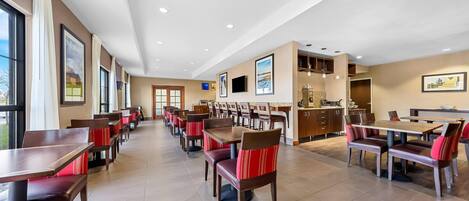 A spacious dining area with multiple tables and chairs, a kitchen area, and a large window providing natural light.