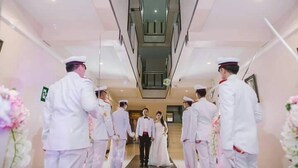 A corridor with a central elevator, flanked by white-clad individuals, leading to a room with a staircase. - Le Parc Wangnang (Phitsanulok)