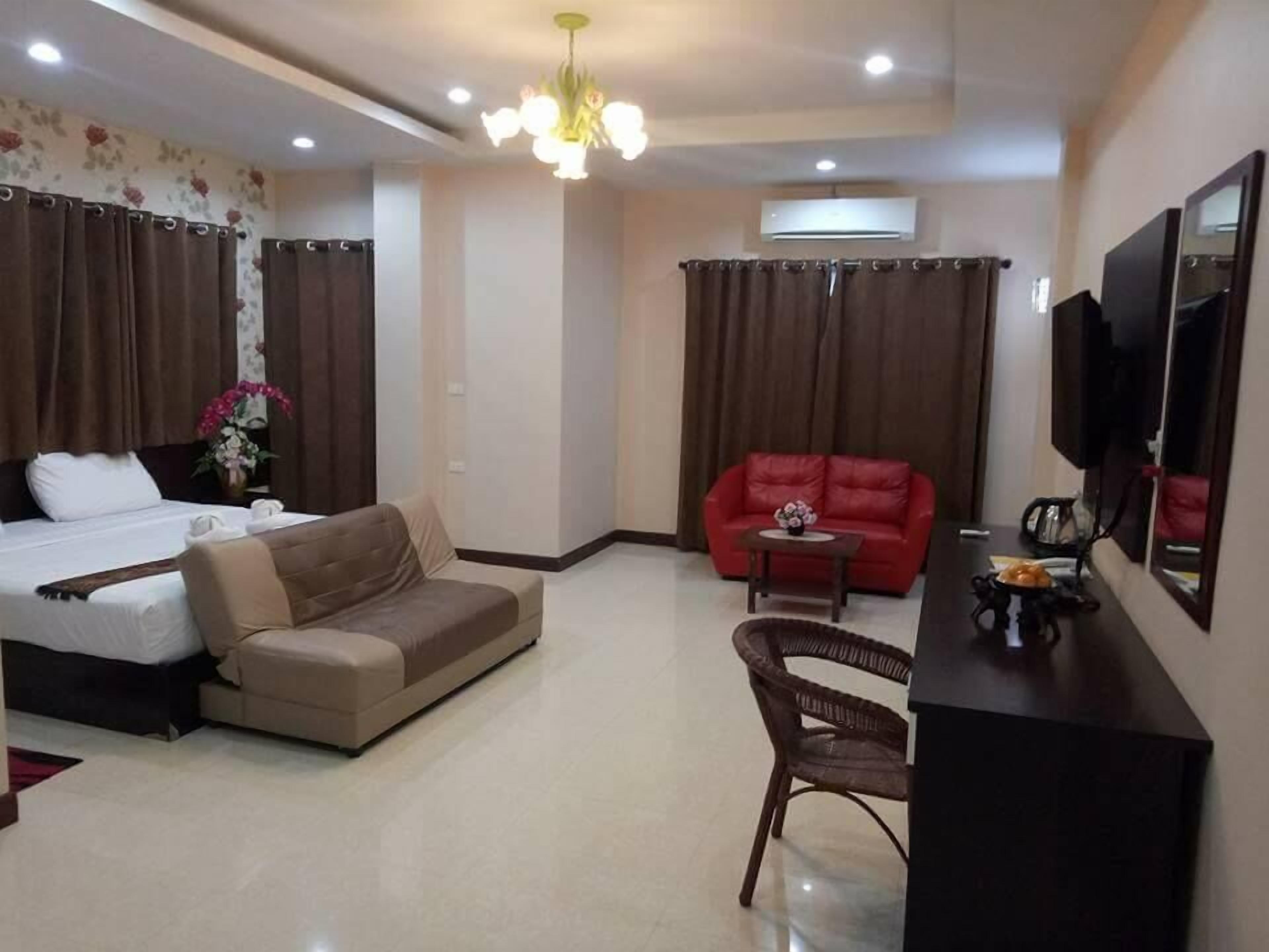A hotel room with a bed, a sofa, a red armchair, a small table, a TV, and an air conditioning unit.