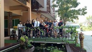 A group of people standing on a balcony with a decorative railing, a small pond with plants, and a statue on the right. - Le Parc Wangnang (Phitsanulok)