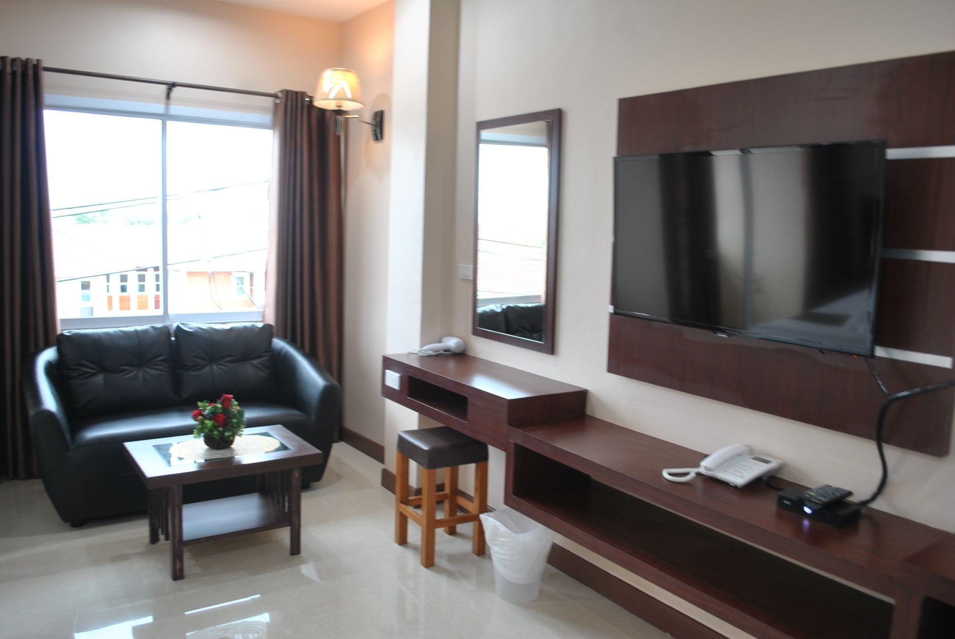 A modern hotel room with a leather sofa, a small table with a vase, a TV mounted on the wall, a desk with a phone, and a trash bin.
