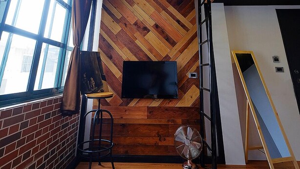 Una habitación con suelo de parqué tipo espiga, pared de ladrillo, un gran ventanal con cortinas, televisor montado, una escalera, un ventilador y una mesa con una silla.