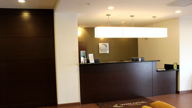 A reception area with a dark wooden reception desk, a welcome mat, and a sign above the desk. - Spark by Hilton Odessa (Odessa)