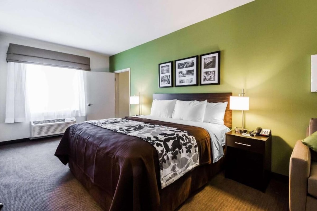 Habitación de hotel con cama, mesitas de noche, lámpara, silla y ventana con cortinas.