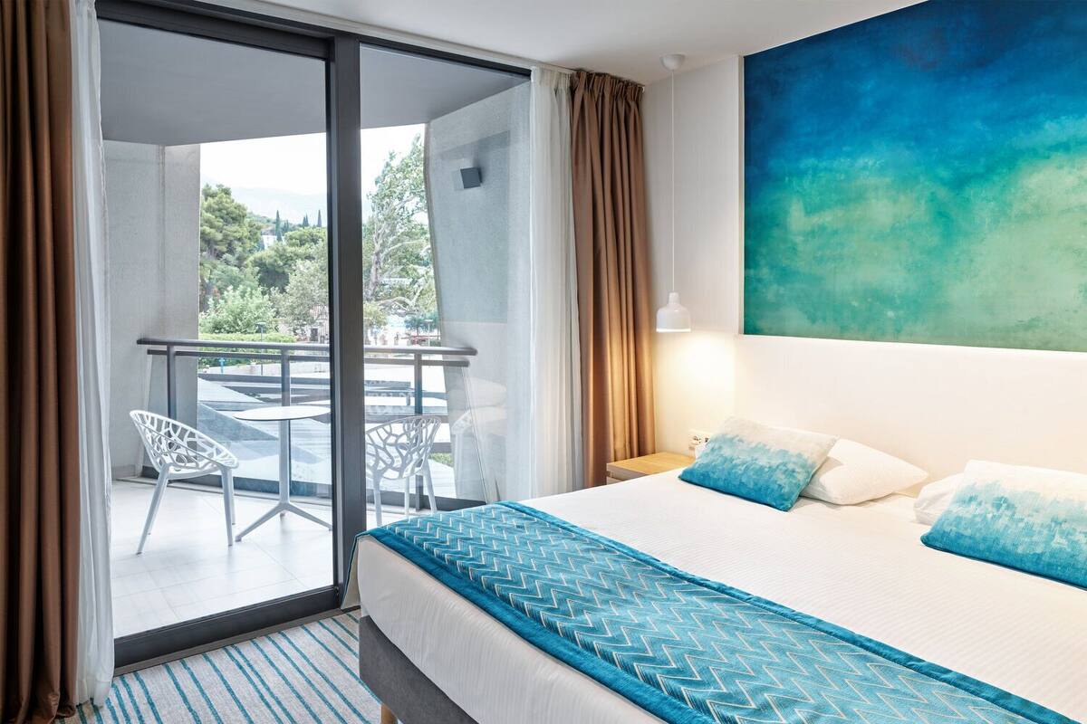 Ein Hotelzimmer mit Bett, Blick ins Freie, einem Balkon mit Stühlen und einem Bild an der Wand.