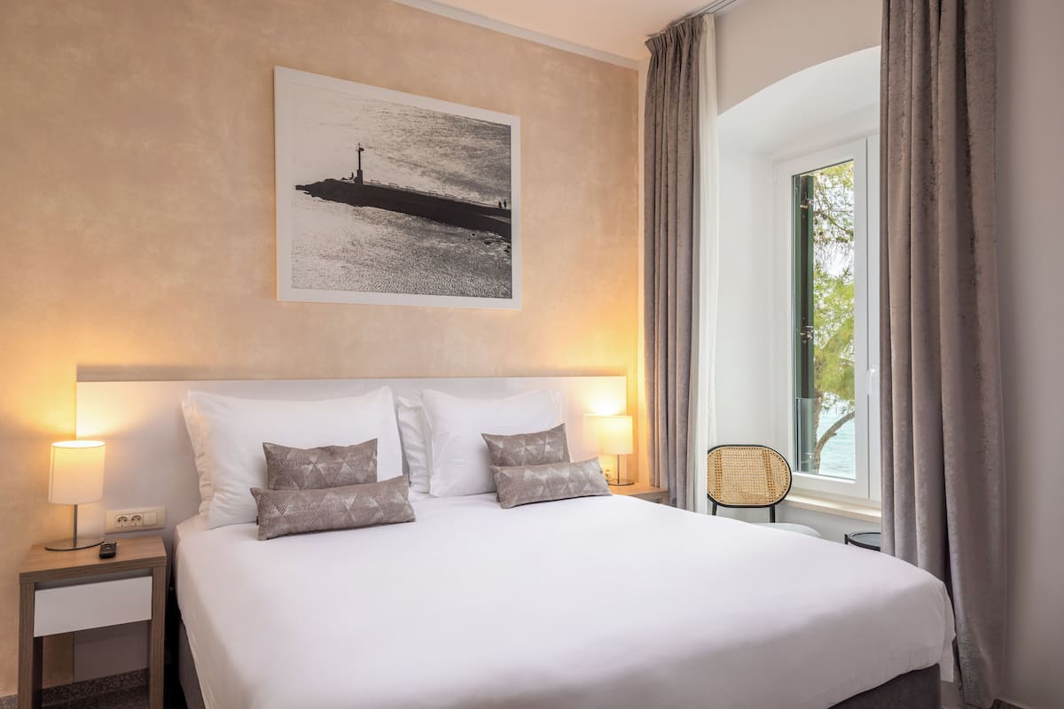 Ein Hotelzimmer mit einem großen Bett, zwei Nachttischlampen, einem gerahmten Bild an der Wand, einem Fenster mit Vorhängen und einem Sessel am Fenster.