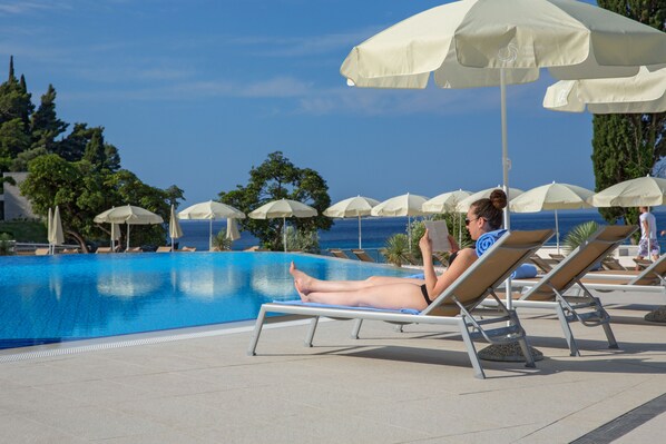 Zona junto a la piscina con sillas reclinables y sombrillas, cielo azul despejado y vista al mar.