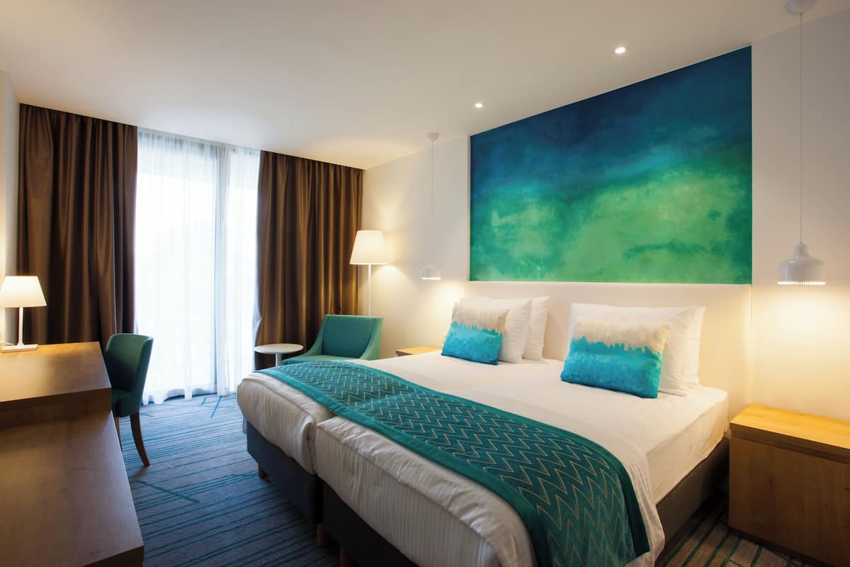 Ein Hotelzimmer mit einem Bett, zwei Sesseln, einem Schreibtisch, einer Lampe und einem großen, abstrakten Gemälde an der Wand.