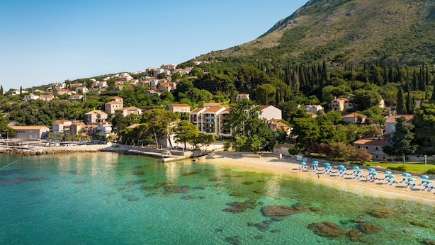 A coastal town with clear turquoise waters, a sandy beach with blue umbrellas, and a backdrop of green hills and residential buildings.