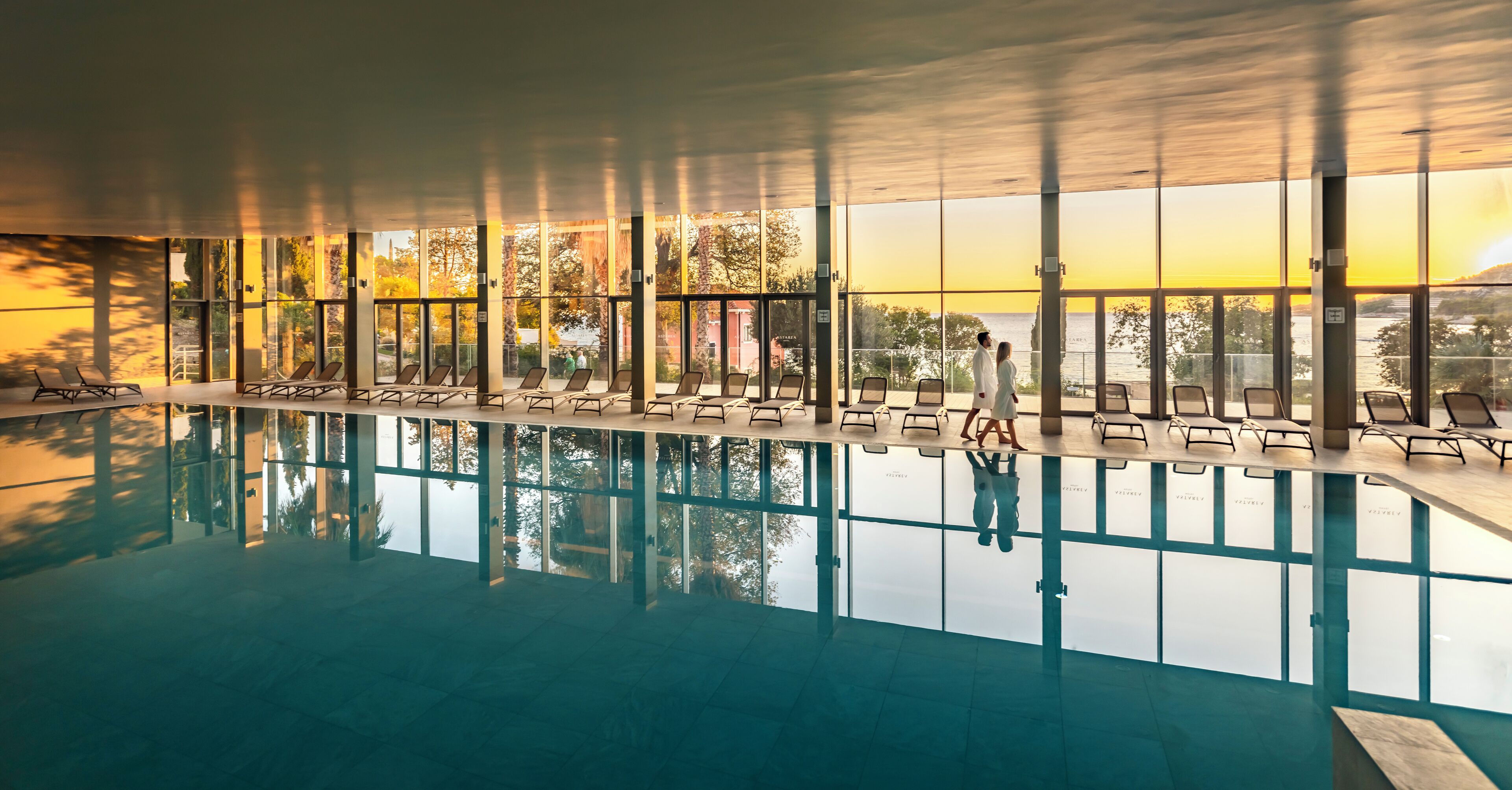 An indoor swimming pool with clear glass walls, lounge chairs, and a view of the sunset.
