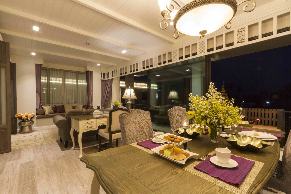 A dining area with a table set for two, a vase with flowers, and a view of the night sky through a large window.