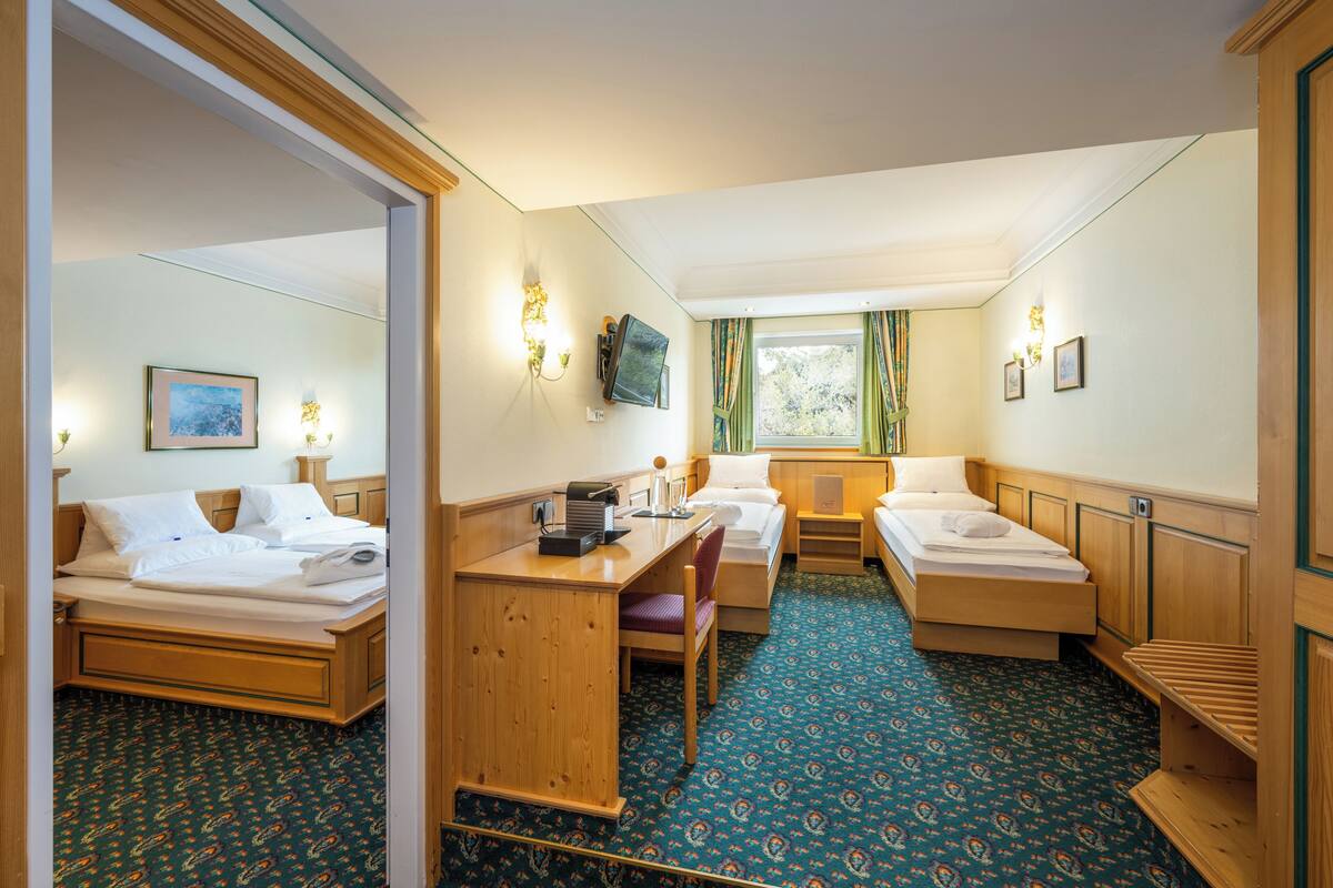 A hotel room with two beds, a desk, and a chair. There is a window with curtains, a flat-screen TV mounted on the wall, and a small table with a lamp.