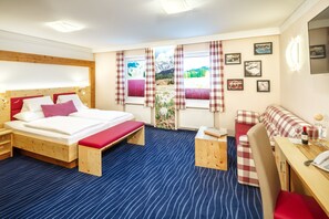 A hotel room with a wooden bed, a small desk, a chair, and a sofa. There are windows with curtains, a plant, and framed pictures on the wall. - Hotel Alpina (Untertauern)