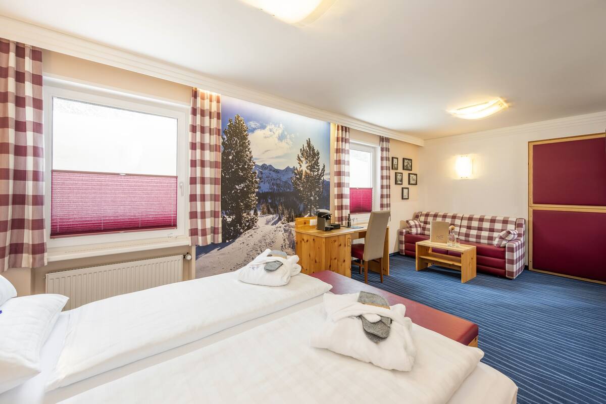 A hotel room with two beds, a desk, a chair, a red sofa, and a large window with curtains.