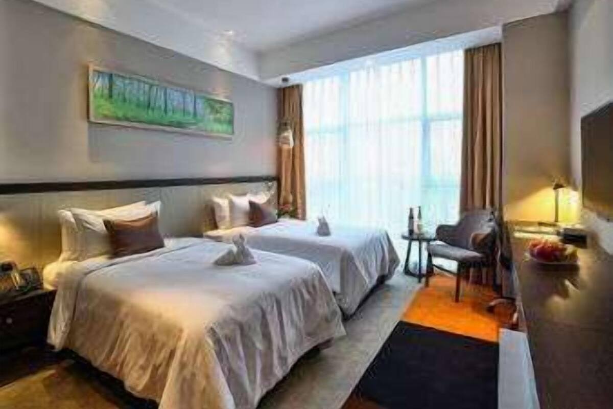 Habitación de hotel con dos camas, un cuadro en la pared, una ventana con cortinas y una mesita con lámpara.
