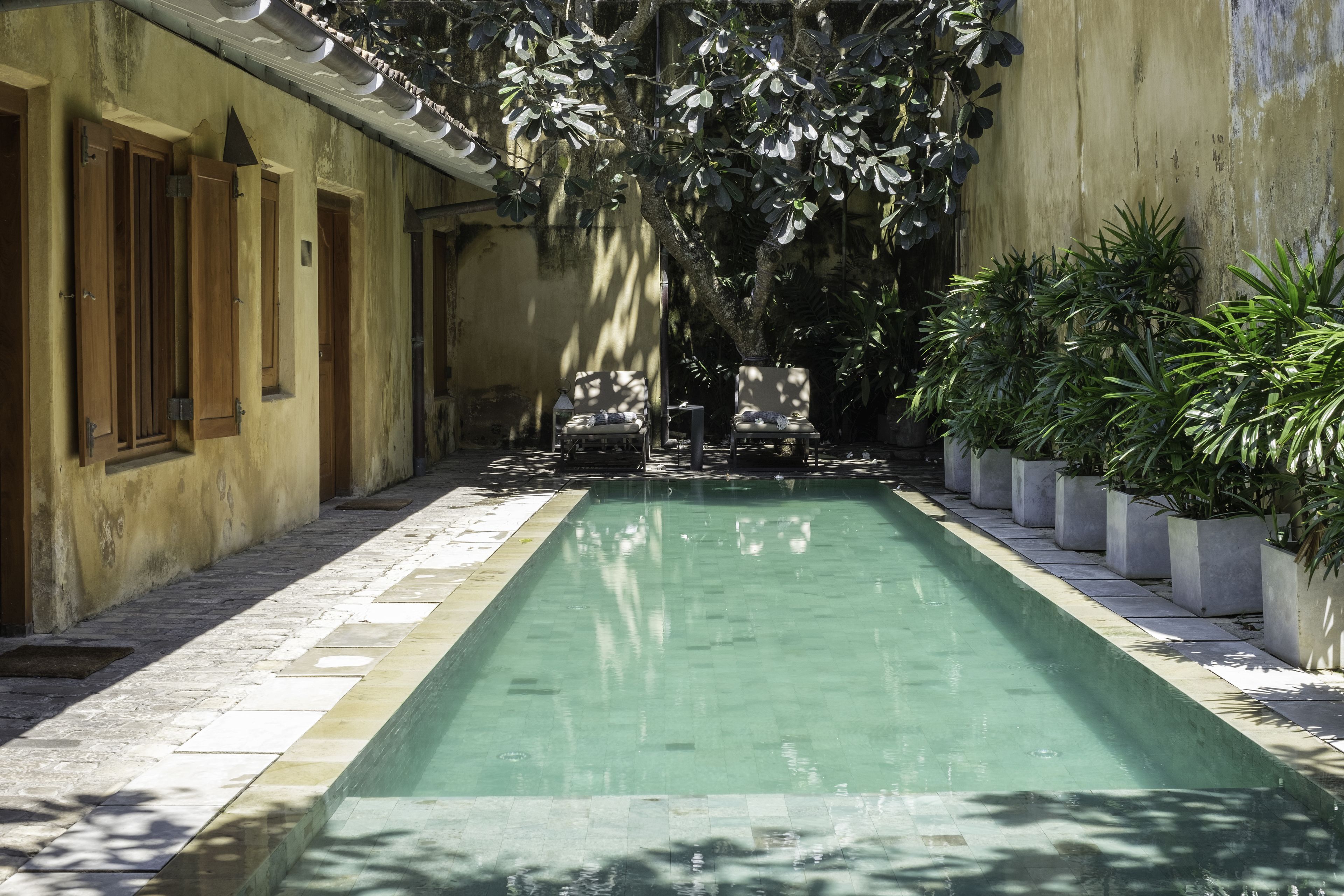 A pool with chairs and potted plants, surrounded by a building with a tiled floor and wooden doors.
