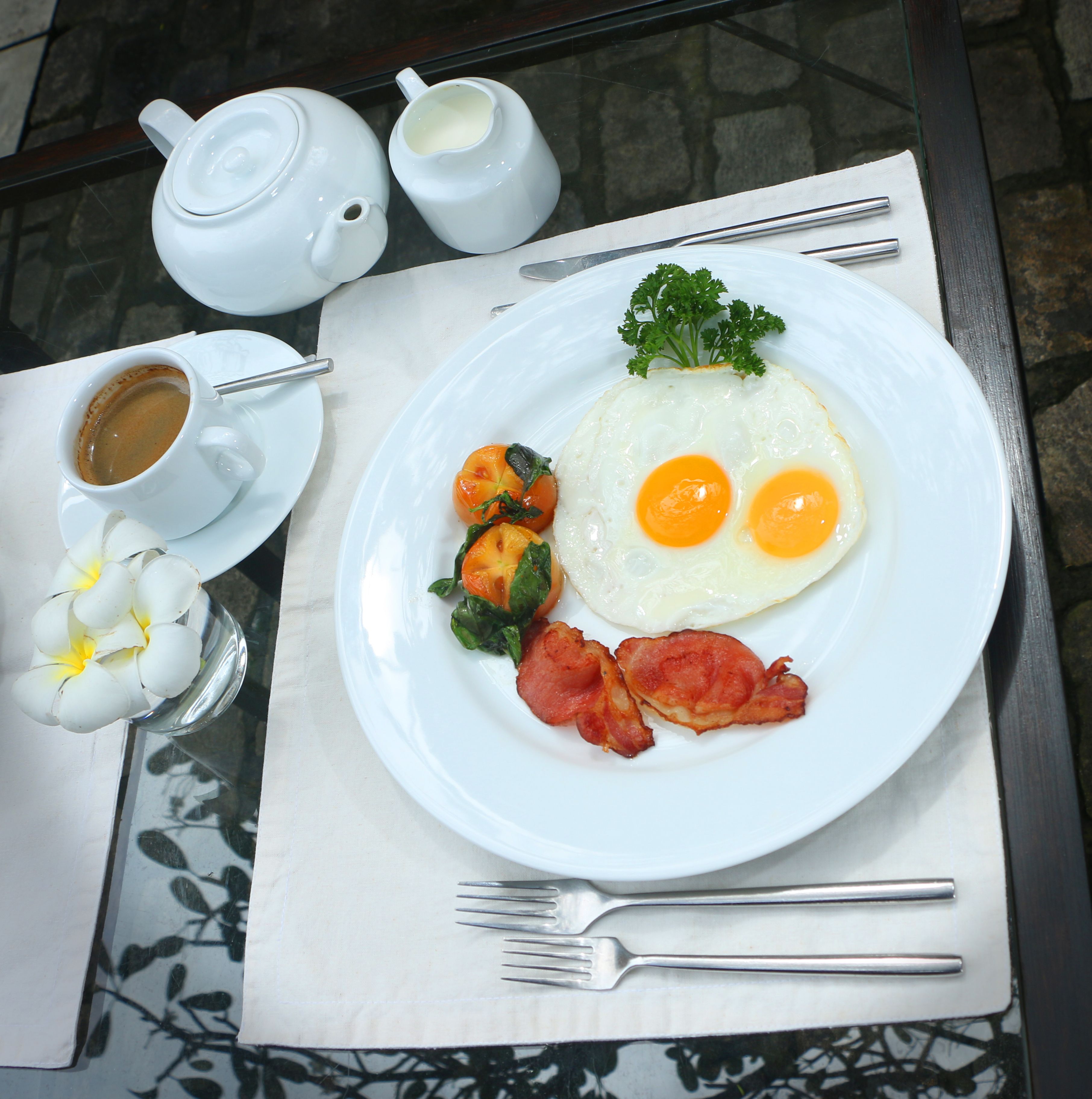 A breakfast plate with eggs, bacon, and tomatoes, accompanied by a cup of coffee and a teapot on a table.
