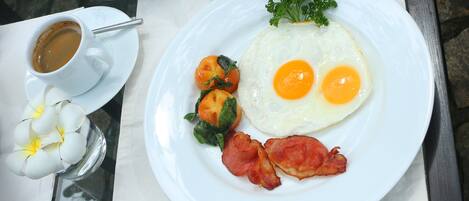 A breakfast plate with eggs, bacon, and tomatoes, accompanied by a cup of coffee and a teapot on a table.
