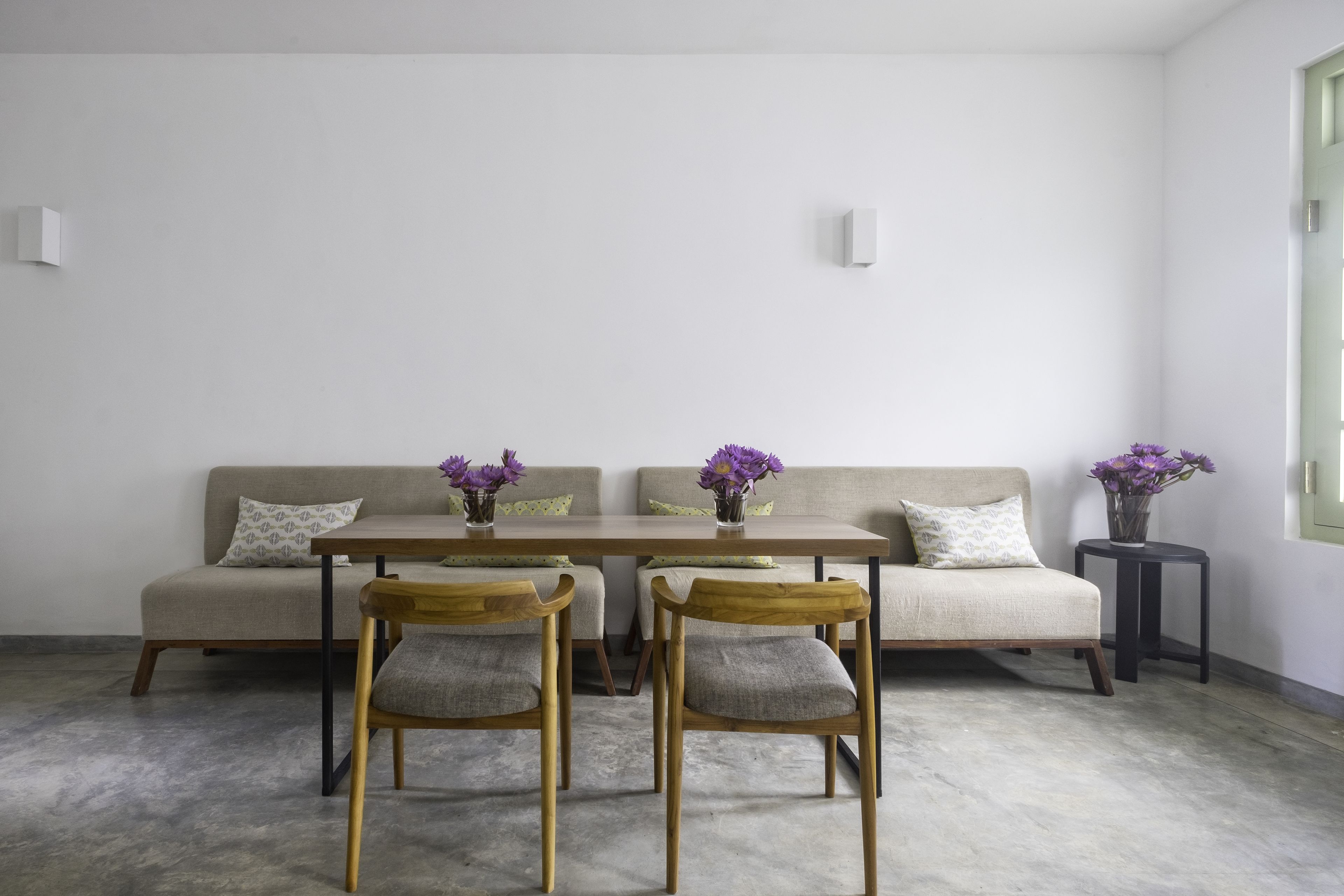 A minimalist living room with a sofa, two wooden chairs, a table, and purple flowers in vases.