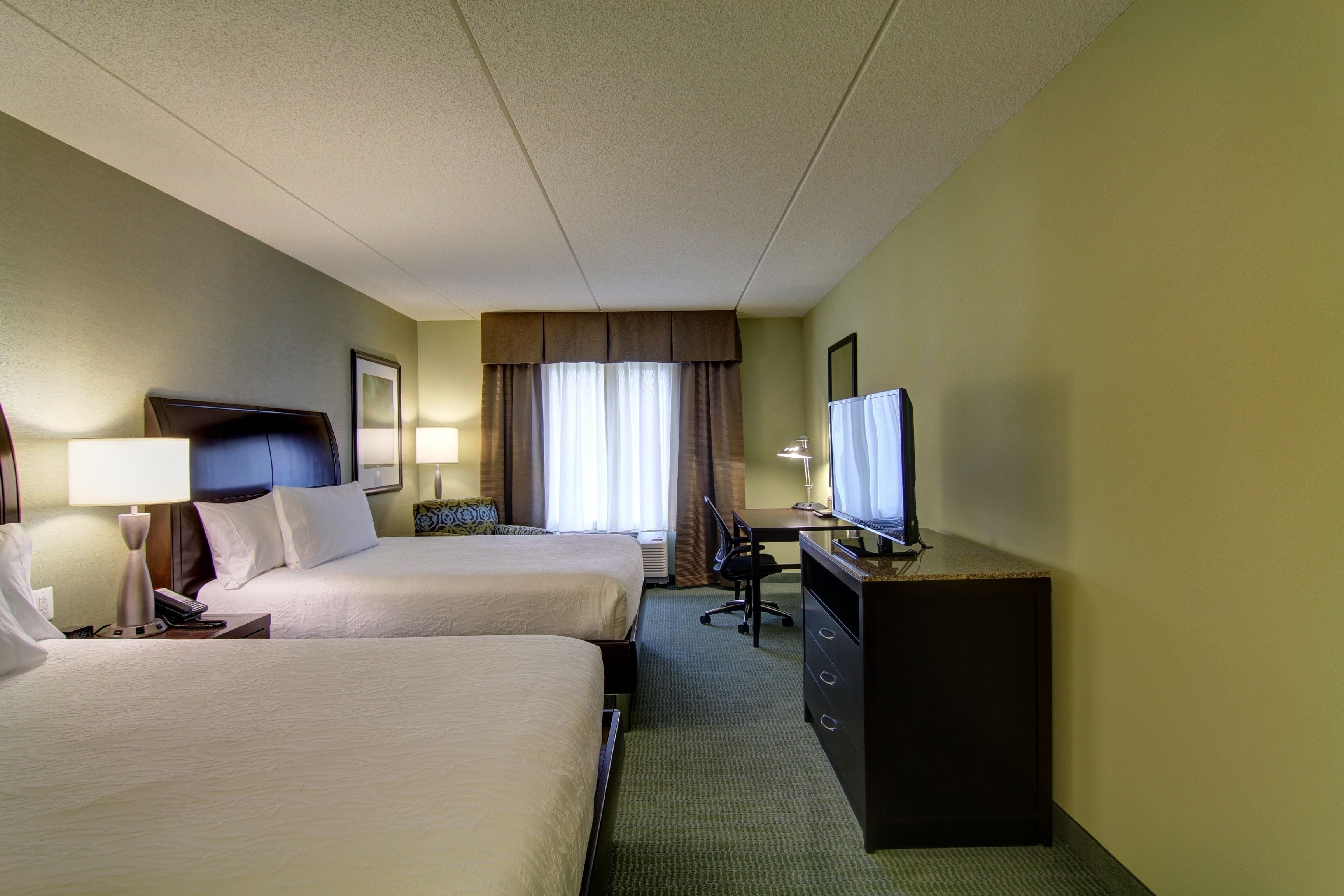A hotel room with two beds, a desk, a television, and a window with curtains.