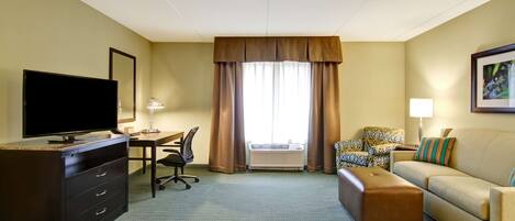 A hotel room with a flat-screen TV, a desk with a chair, a sofa, an ottoman, and a large window with curtains.