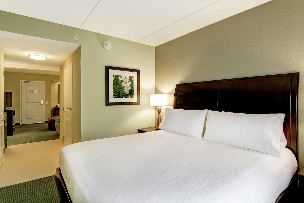 Une chambre d’hôtel avec un grand lit, une table de chevet avec une lampe, un tableau encadré au mur et une porte donnant sur une salle de bain.