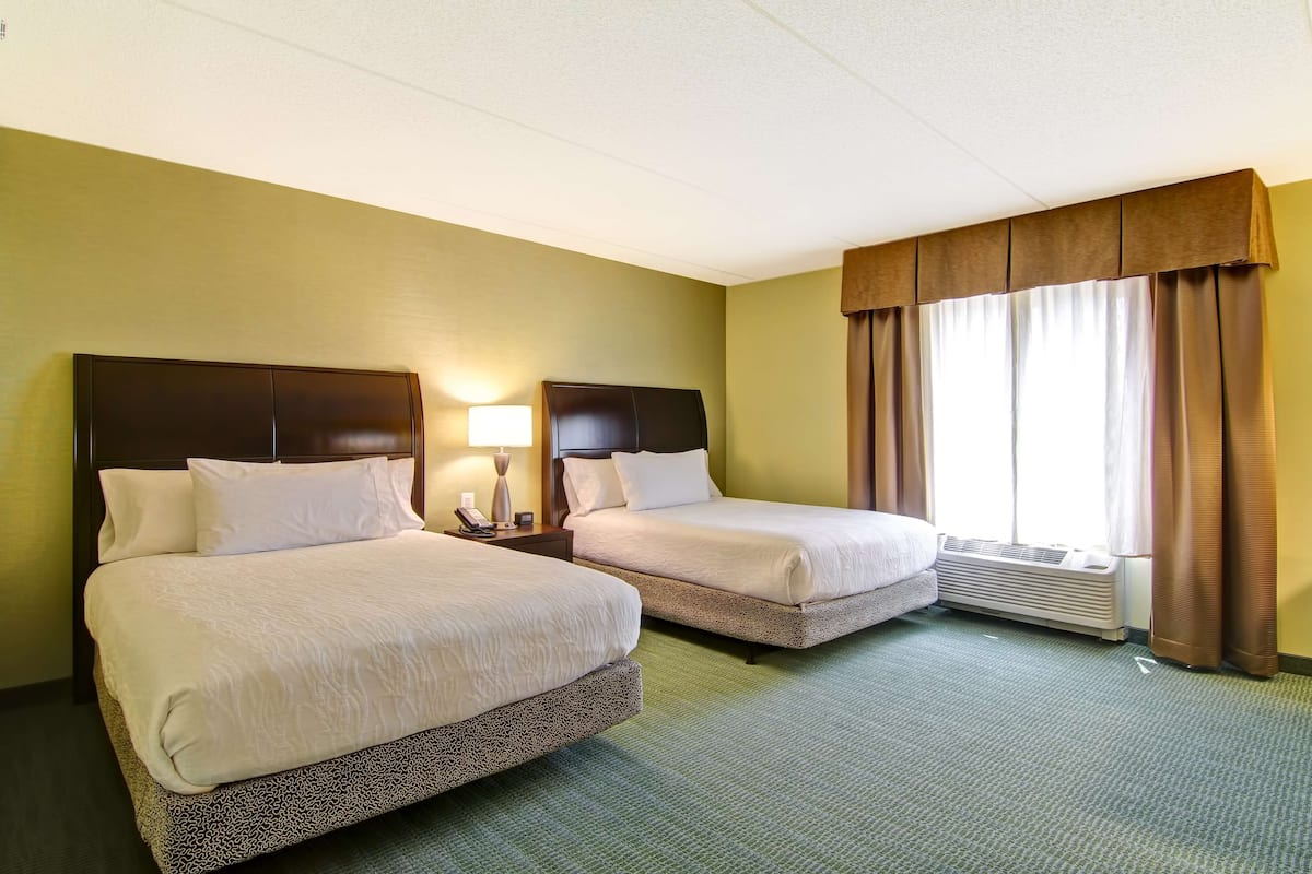 Une chambre d’hôtel avec deux lits, une table de chevet avec une lampe, une fenêtre avec des rideaux et un climatiseur fixé au mur.
