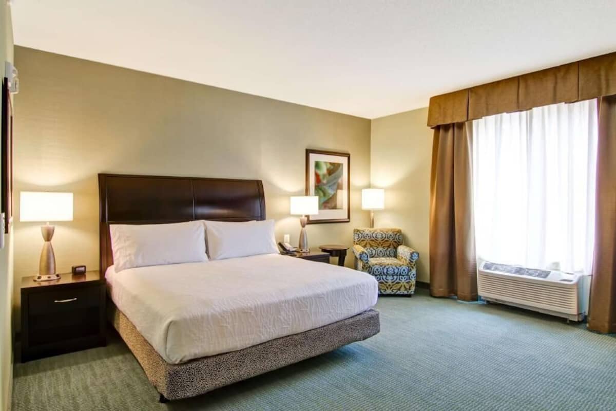 Une chambre d’hôtel avec un grand lit, deux tables de chevet avec des lampes, une chaise, un tableau encadré au mur et un appareil de climatisation.