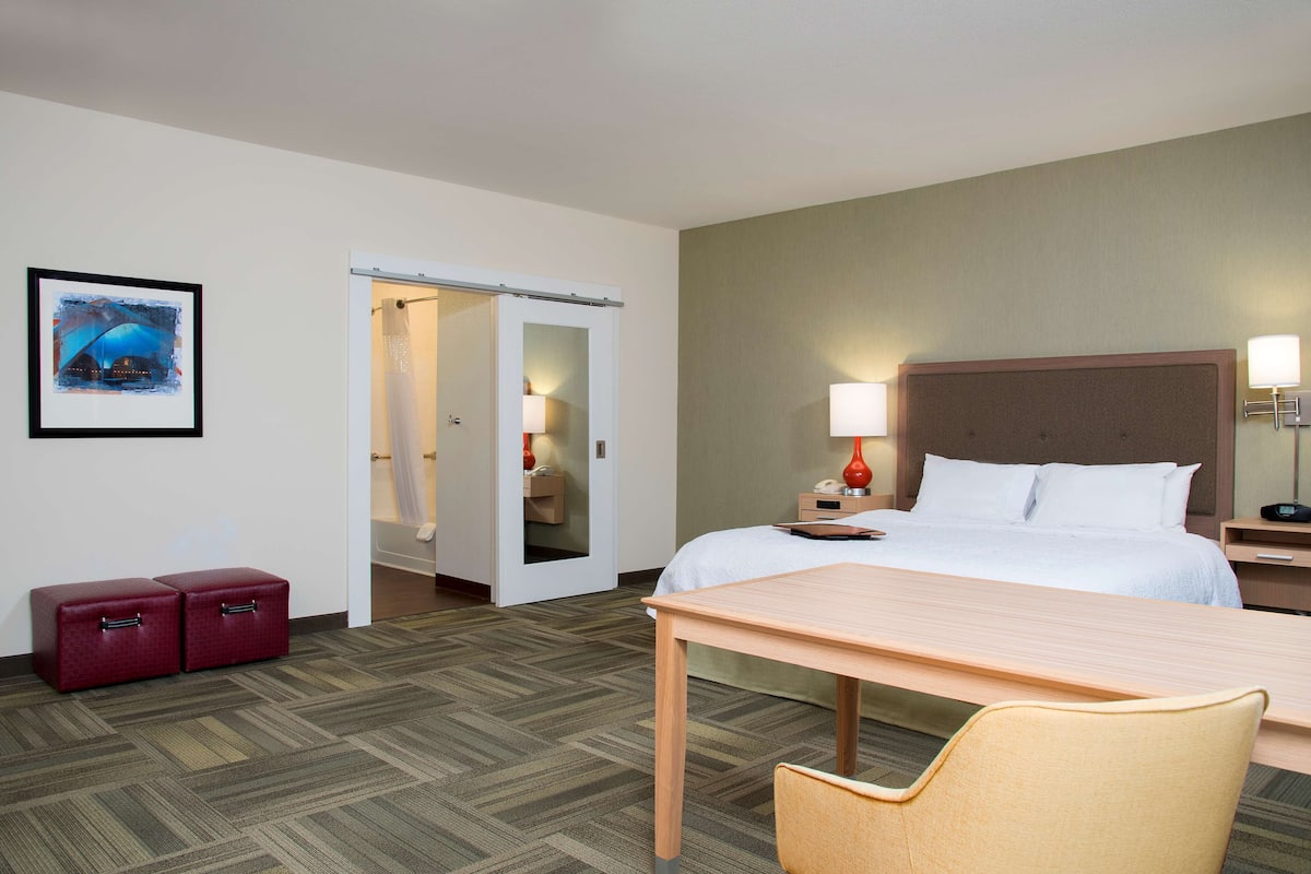 A hotel room with a bed, bedside table, lamp, and a chair. There is a door leading to another room and a small red cabinet.