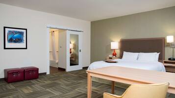A hotel room with a bed, bedside table, lamp, and a chair. There is a door leading to another room and a small red cabinet.