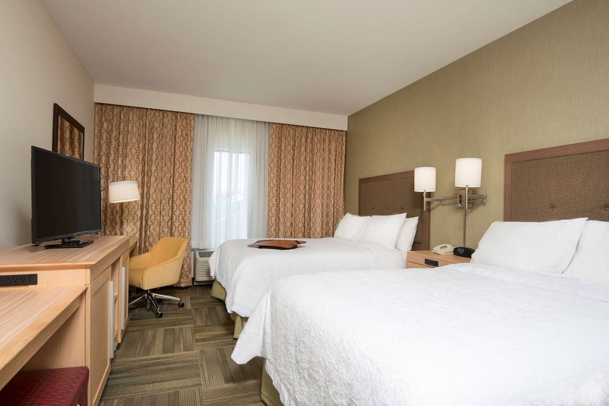 A hotel room with two beds, a desk with a TV, a chair, and a lamp.
