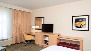 A hotel room with a bed, a desk with a computer, a chair, a TV, a red bench, and a framed picture on the wall.