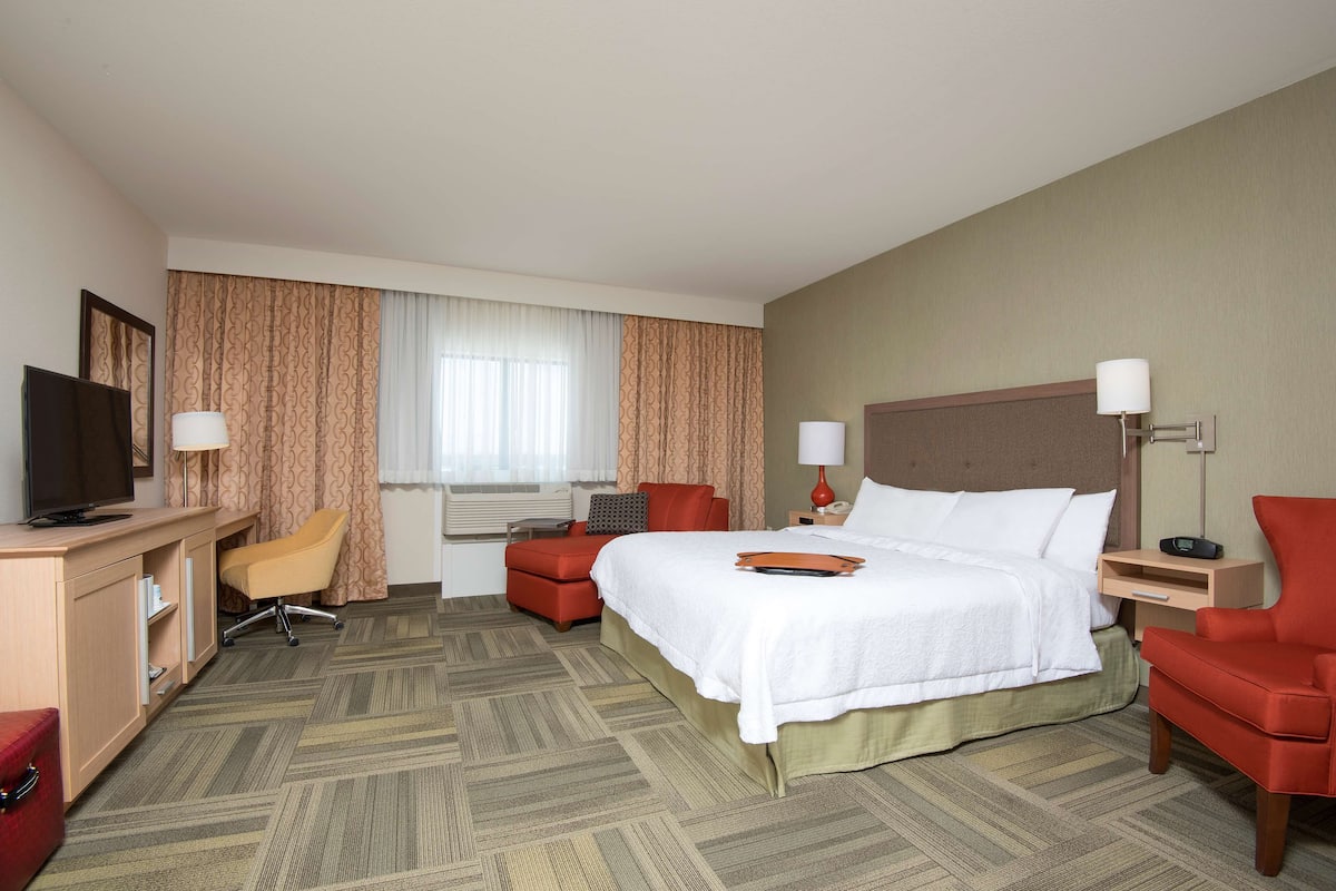 A hotel room with a bed, a desk with a TV, a chair, a red armchair, and a nightstand with a lamp.