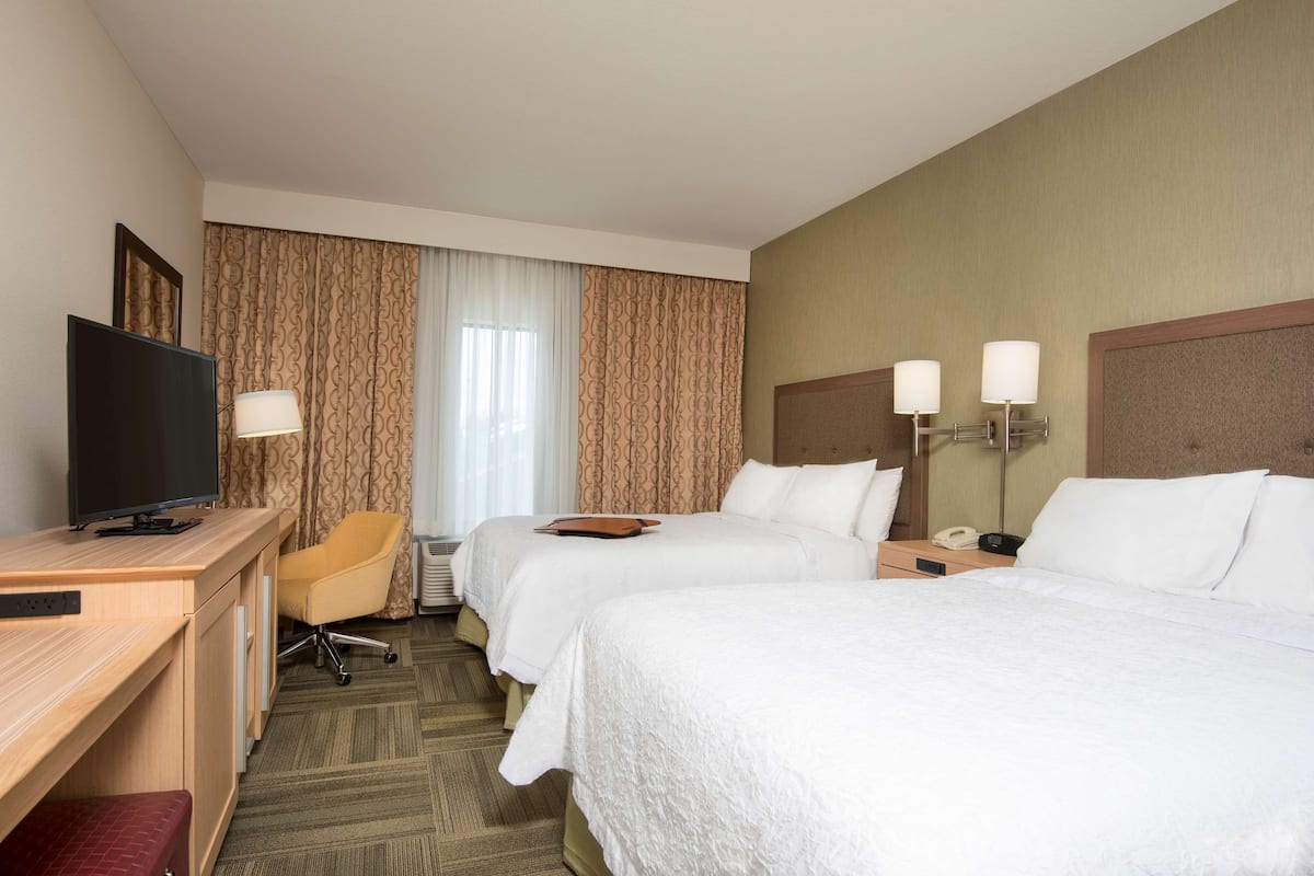 A hotel room with two beds, a desk with a television, a chair, and a bedside table with lamps.