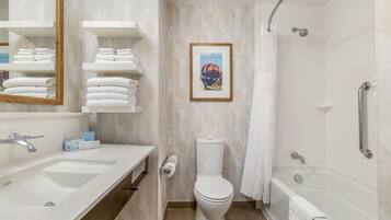 Un baño con lavamanos, inodoro y ducha. Hay toallas en la pared y un cuadro enmarcado sobre el inodoro.