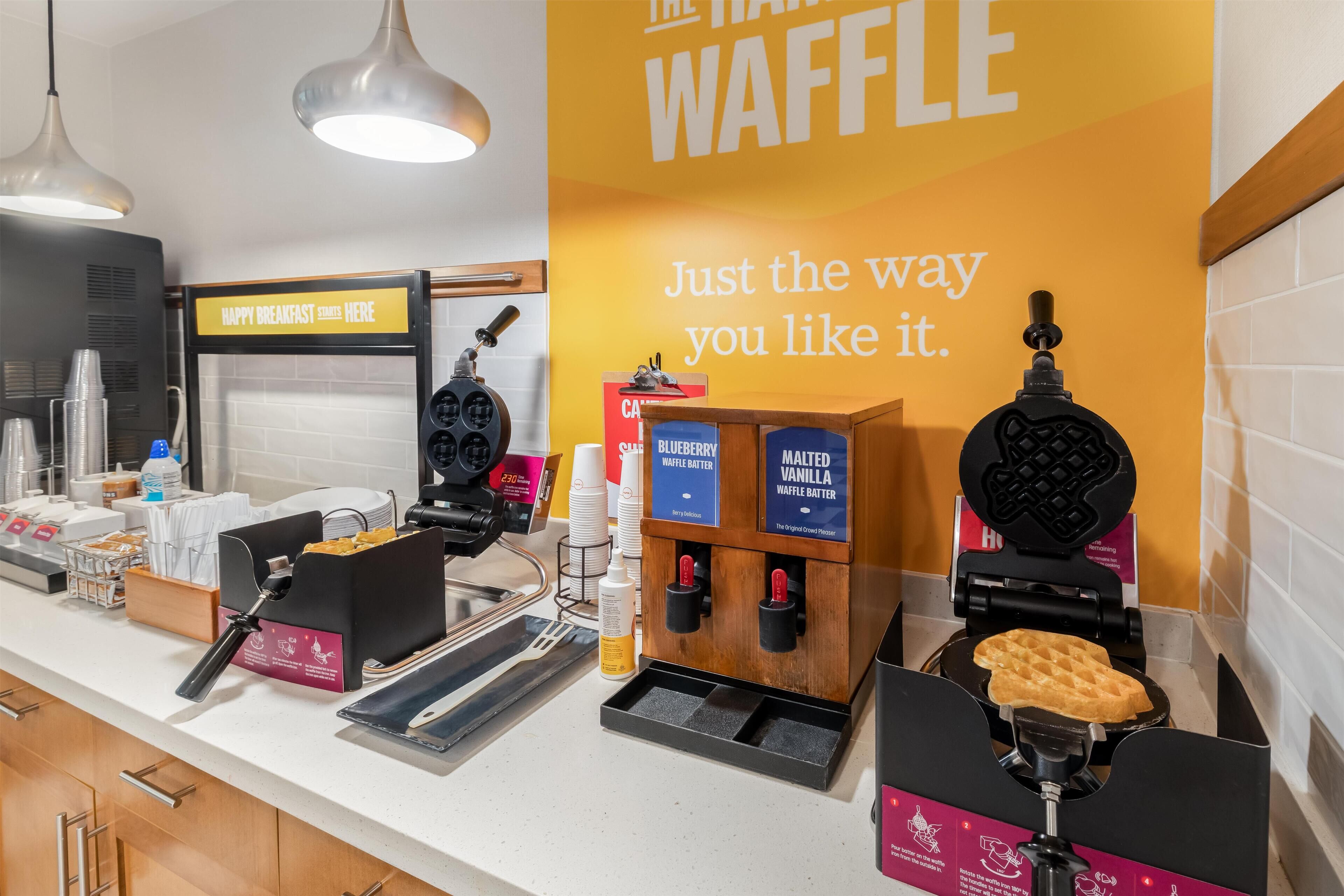 A waffle station with options for blueberry and vanilla waffles, a coffee machine, and a sign indicating a happy breakfast.