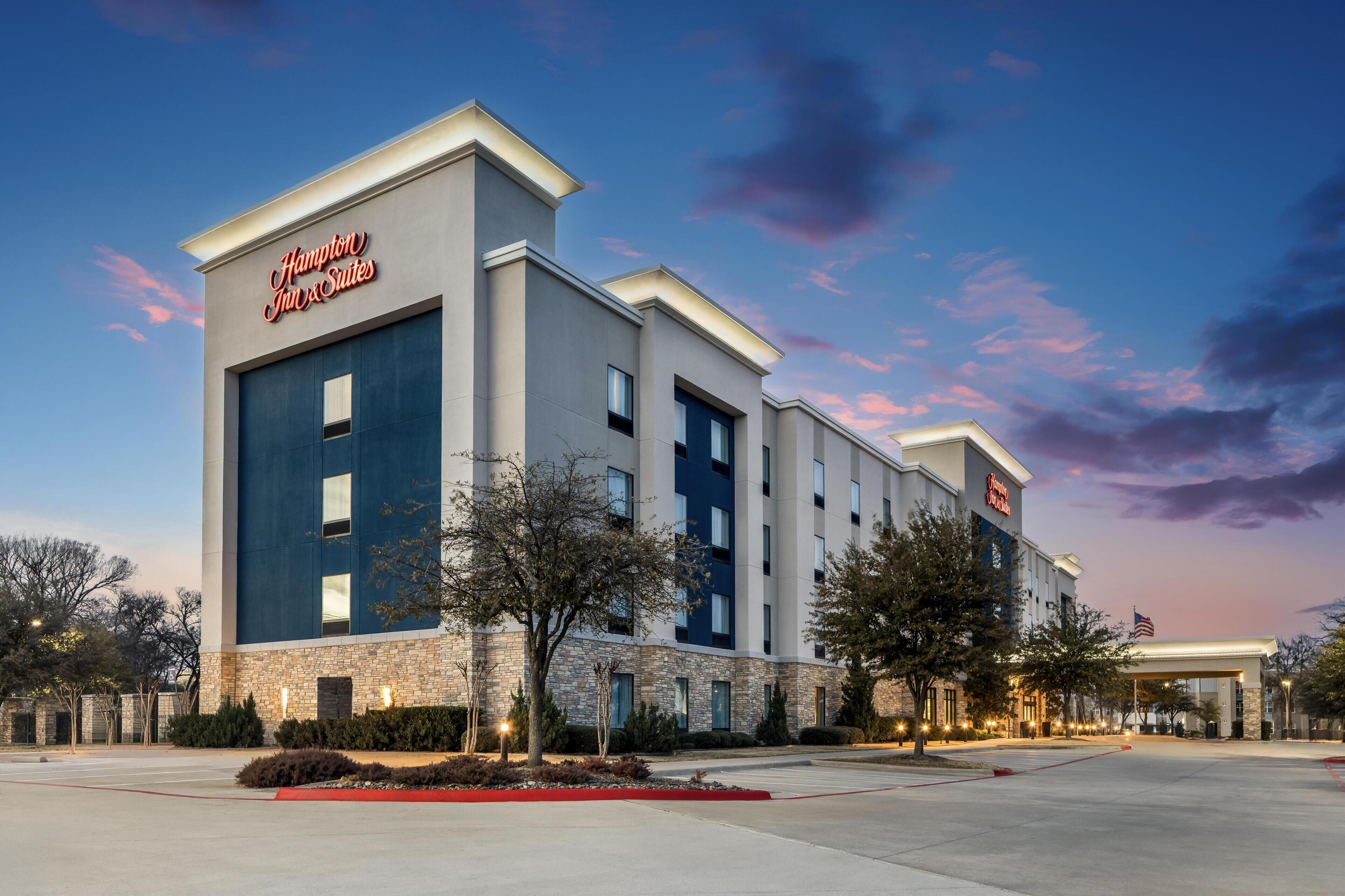 Et Hampton Inn & Suites hotel med et moderne design, karakteriseret ved en stenfassade, mange vinduer og et flag.