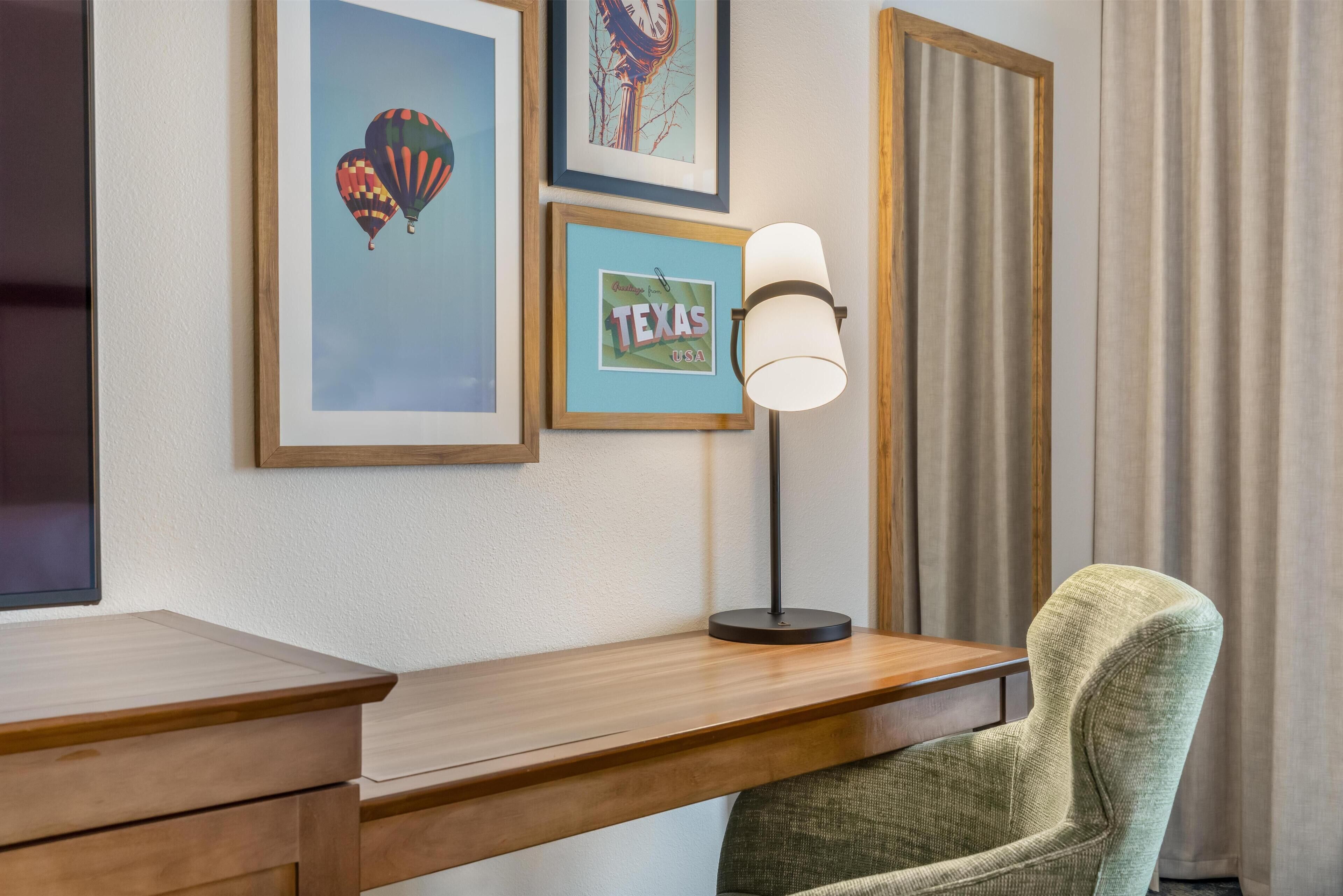 A wooden desk with a lamp, a green chair, and framed pictures on the wall.