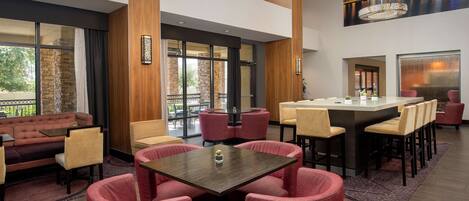 A modern hotel lobby with a seating area, a bar, and large windows offering views of the outdoors.