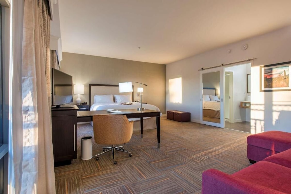A hotel room with a bed, desk, chair, and a mirror.