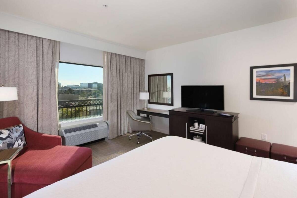 Une chambre d’hôtel comprenant un lit, un bureau avec un ordinateur, une télévision, une chaise, un canapé, une fenêtre avec des rideaux et une vue sur la ville.