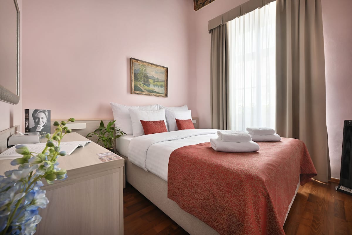 A hotel room with a bed, a desk, a chair, a plant, and a painting on the wall.