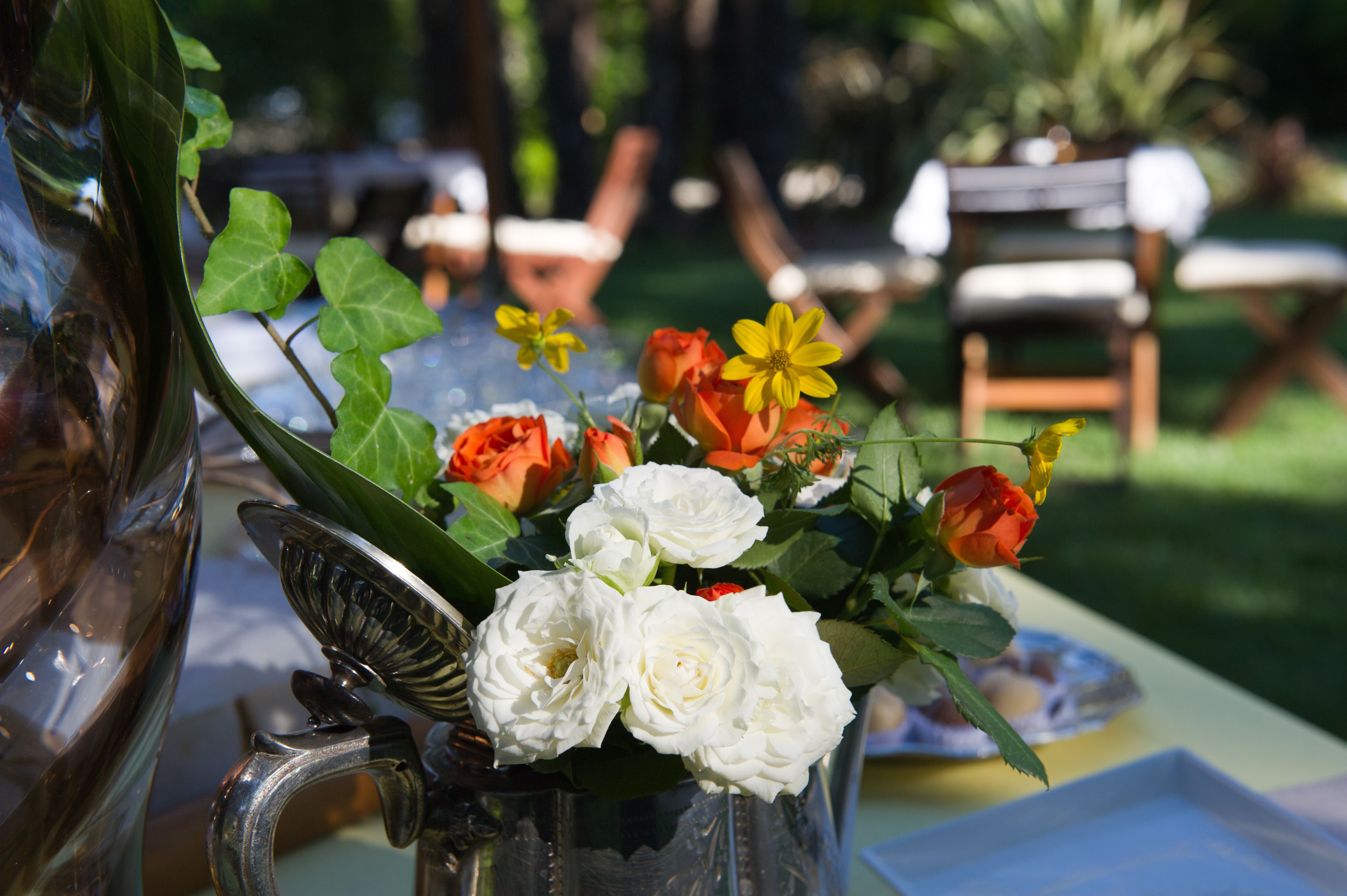 Una mesa con una jarra metálica que contiene flores blancas y anaranjadas, un florero de vidrio y un plato con comida.