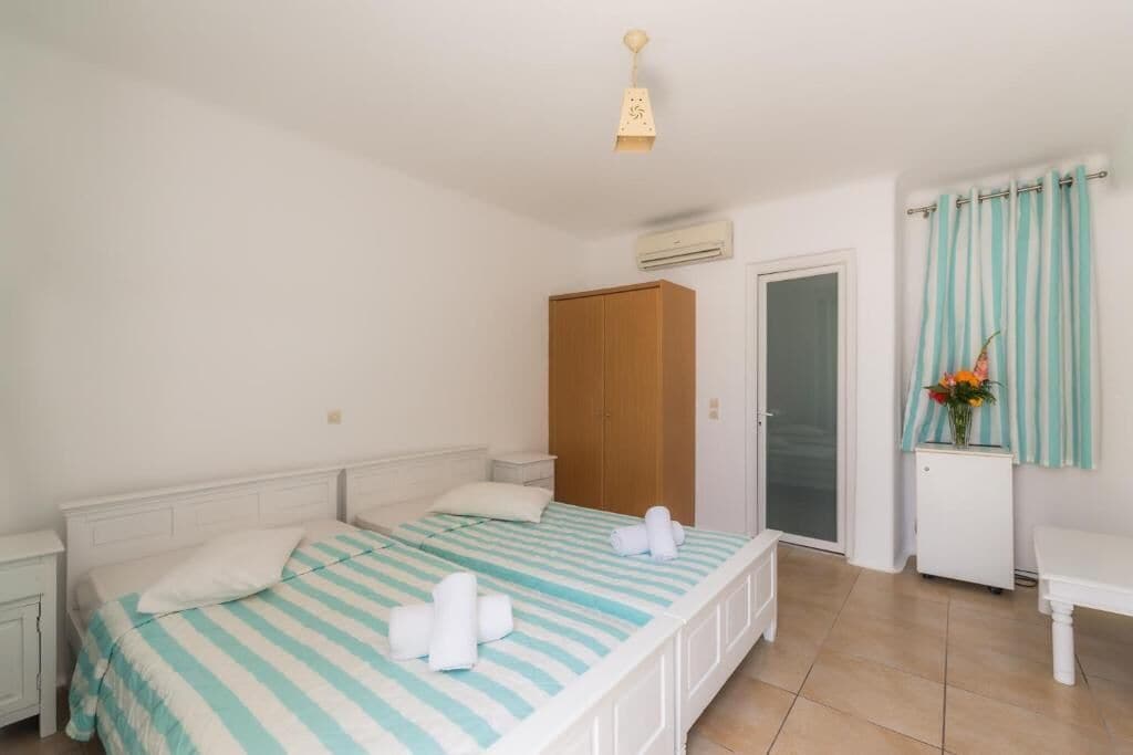 A bedroom with a bed, a wardrobe, an air conditioning unit, a small table with a vase of flowers, and a curtain.