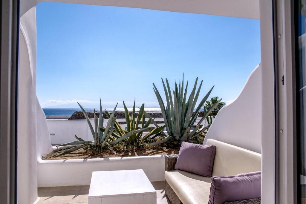 A balcony with a white sofa and a small table, overlooking a beach with agave plants and a clear sky.