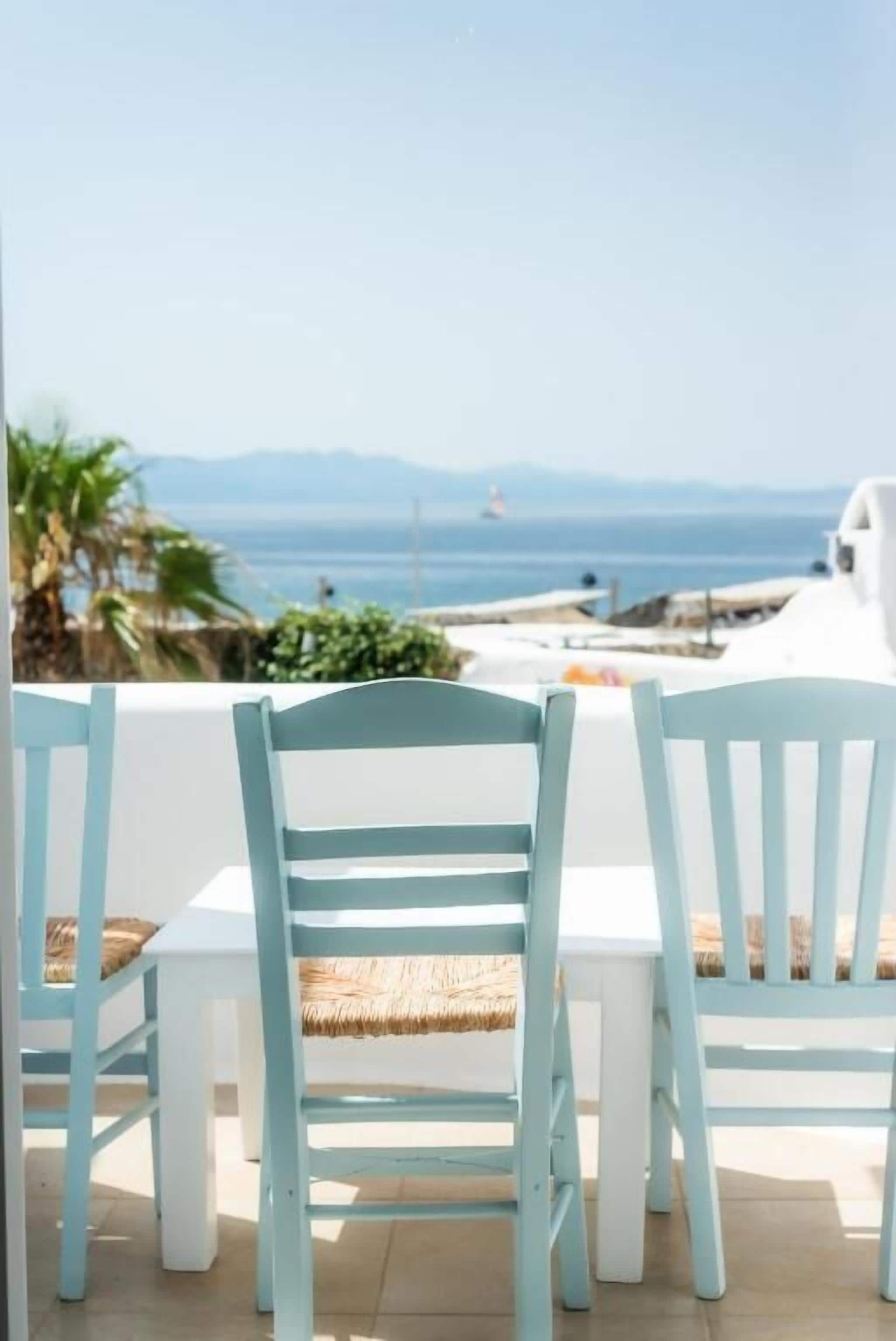 A seaside patio with light blue chairs and a white table, overlooking the ocean and a white building.