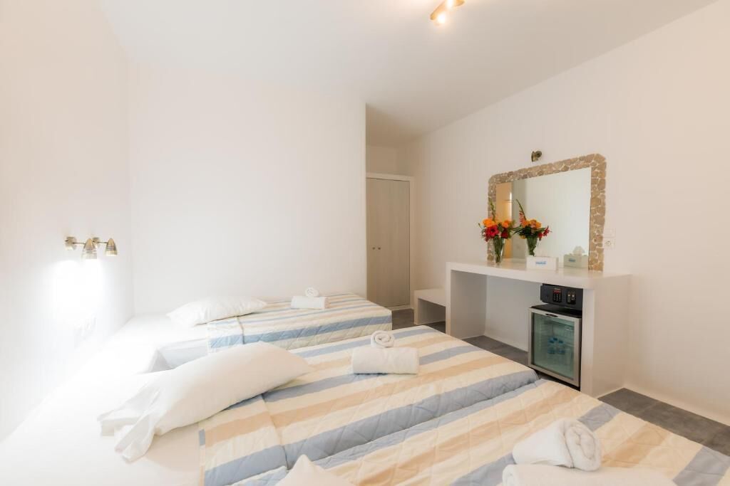 A hotel room with two beds, a small kitchenette, a mirror, and a vase with flowers.
