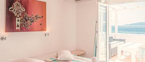 A bedroom with two beds, a painting on the wall, and a view of the sea through an open door.