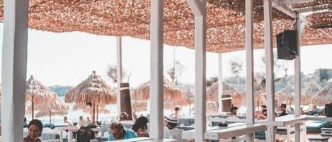 An outdoor dining area with white wooden tables and chairs, covered by a thatched roof.