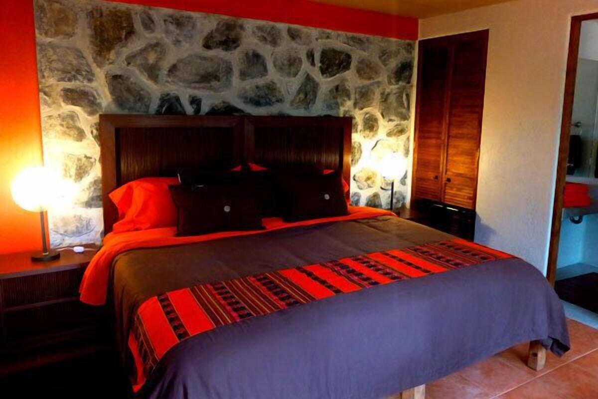 A bed with a stone wall backdrop, wooden headboard, and a red and black bedspread.